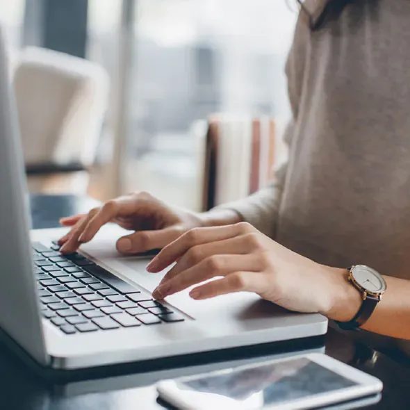 Hands at a laptop computer with a watch on the left wrist and a mobile phone on the table