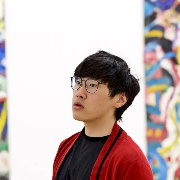 Asian man in glasses and red cardigan standing in front of a wall with art on it
